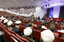 A projection from above into the auditorium, many people, some of them wearing white caps on their heads. A lectern in the middle of the auditorium, a person stands behind it. Behind it a grey curtain and a decoration in blue and white.