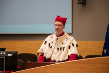 A middle-aged man, dressed in a red and white toga, with a red biretta on his head. He speaks into a microphone.