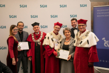 A group of people stand and smile for the camera. Some of them are dressed in academic togas.