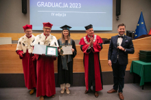 A group of people standing in the middle of the room. Some of them dressed in academic togas. The man on the right, dressed in a suit, holds a microphone in his hands and speaks.