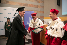 In the main foreground, three people dressed in academic togas. Two of them are middle-aged men in red and white togas, one man wearing a black toga.