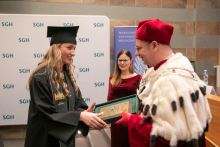 Three people, two women and one man. A man wearing a red and white academic toga hands a diploma to a woman wearing a black toga.