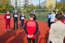 Grupa osób ubranych w stroje sportowe. Stoją na boisku sportowym, w tle bramka do piłki nożnej.