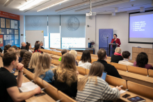 Sala wykładowa. Na pierwszym planie ławki i słuchacze widoczni tyłem. Na końcu sali kobieta wygłasza wykład, a za nią ekran.