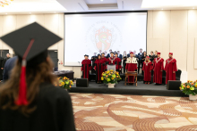 In the foreground, a group of people standing on an elevation. The individuals are dressed in academic attire, behind them a multimedia screen with a red and black logo.