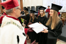 A group of people dressed in black academic togas and a man dressed in a white-red toga handing one person an open-covered diploma.