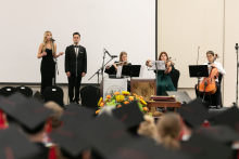 A few people on a raised platform. Women and man stand at microphones and sing, three people sit and play instruments.