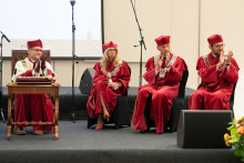 Four people, seated. They are dressed in red academic togas. They are applauding.