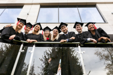 A group of young people dressed in academic togas pose on a glass balcony.