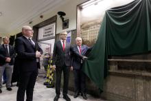 Na zdjęciu kilka osób, stoją przed tablicą powieszoną na ścianie. Dwóch mężczyzn stojących pod tablicą trzyma zielone sukno.