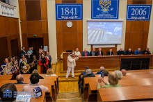 Aula. Za stołem prezydialnym siedzą mężczyźni w garniturach. Widać osoby w strojach ludowych i publiczność.