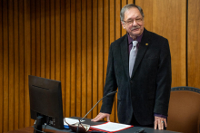 An older man in a suit stands at a desk.