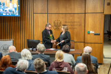 Lecture hall full of audience. At the presidential table, a woman presents flowers to an elderly man.