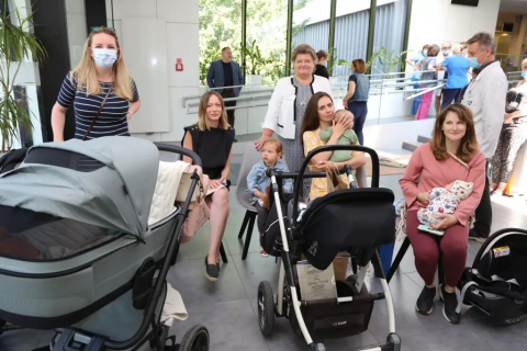 Hol szpitala. Na pierwszym planie od lewej młoda kobieta z długimi włosami siedzi z dzieckiem na kolanach. Obok pośrodku kolejna długowłosa kobieta z niemowlakiem na rękach i kilkulatkiem siedzącym obok, przed nimi wózek-gondola . Z prawej kolejny wózek-gondola za nim dwie kobiety. Jedna w bluzce w paski - stoi, druga w czarnej bluzce  - siedzi. Za nimi pośrodku stoi starsza kobieta w białym żakiecie. W tle stoją i rozmawiają ludzie - niektórzy w fartuchach lekarskich i maseczkach chirurgicznych. 