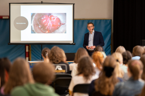 Lecture hall. In the foreground, heads of the audience visible from behind. At the end of the room a man in a suit and a headset microphone. In his hand he holds a remote control. Behind him, the screen shows pictures of a human liver.