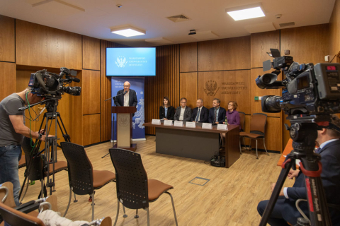 Sala konferencyjna. Z prawej i z lewej strony kamery telewizyjne. w głębi mównica, przy której stoi mężczyzna w garniturze. Obok mównicy stół, przy którym siedzą dwie kobiety i trzech mężczyzn.