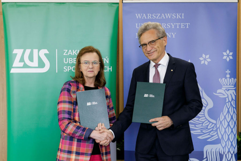 On the left, a woman wearing glasses and a checked jacket. On the right, a man in glasses and a suit. They stand and shake hands, looking into the camera. In their hands they hold green briefcases.