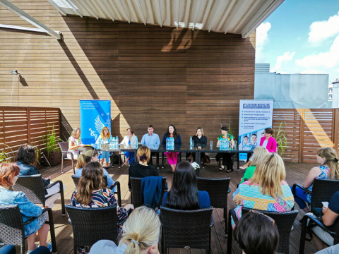 A group of people sitting behind a table, facing the camera. Behind them a wooden wall and two rollups. With their backs to the camera - listeners sitting on chairs