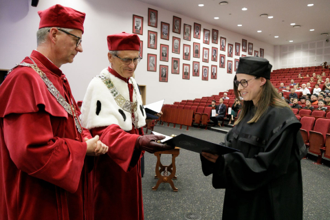 Two men and a woman in togas in the auditorium. A man in a red toga hands a diploma to a woman in a black toga.