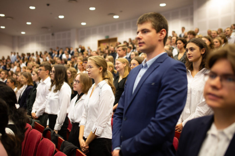 Grupa młodych ludzi ubranych elegancko stoi na auli w rzędach czerwonych krzeseł.