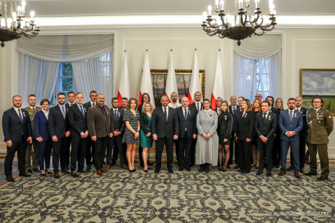 A group of people posing for a photo in a large room. They are all standing. They are formally dressed. Behind them are the flags of Poland.