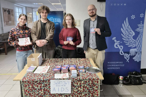 Grupa czterech osób, studentów WUM, stoją i trzymają w dłoniach materiały dotyczące AIDS, po prawej niebieski rollup z logo WUM