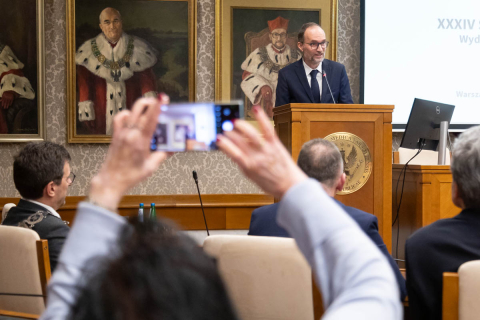 Odkrycie mikroRNA tematem XXXIV Sesji Noblowskiej. Elegancko ubrany mężczyzna stoi za mównicą i przemawia. Osoba trzyma w rękach uniesiony telefon komórkowy i robi mu zdjęcie.